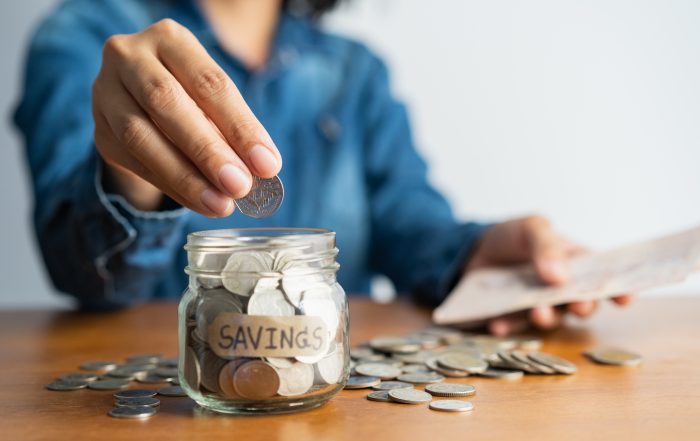 The woman hand is putting a coin in a glass bottle and a pile of coins on a brown wooden table,Investment business, retirement, finance and saving money for future concept.